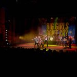 Wide-angle shot of a live stage performance of Big Girls Don't Cry, featuring a group of male vocalists in coordinated grey blazers and red ties mid-choreography. A full backing band with drums, keyboard, and guitars supports them against a brightly lit backdrop emblazoned with the show's title. Colourful stage lighting and a darkened audience add to the theatrical atmosphere.