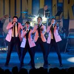 Four male vocalists perform energetically at the front of a stage, dressed in matching pink blazers, black trousers, black ties, and white shirts. Each singer holds a microphone and gestures toward the audience, backed by a live band featuring a bassist, drummer, and keyboardist. The illuminated drum kit and dramatic lighting enhance the lively atmosphere, with a backdrop reading BIG GIRLS DON'T CRY behind them.