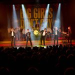 A live stage performance of Big Girls Don’t Cry shows four male singers in black leather jackets, white shirts, and black trousers engaging the audience under dramatic spotlighting. Behind them, a full band plays against a large backdrop bearing the show’s title. The packed theatre is dimly lit, drawing focus to the energetic performance under warm and vibrant stage lights.