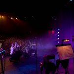 Backstage view of a live performance showing five male performers kneeling dramatically at the front of the stage, dressed in white shirts with black braces and ties. They reach toward the audience under purple and amber stage lighting. The packed theatre crowd watches attentively. In the foreground, a keyboard setup, microphone stands, and a music stand with visible sheet music add depth to the theatrical scene.