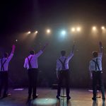 A striking backstage photo captures four male performers standing in a row, backs to the camera, waving to a darkened audience under bright stage lights. Dressed in white shirts with black braces and black trousers, they stand in a misty, atmospheric glow, their silhouettes framed by the warm amber and cool blue spotlights. The image evokes the closing moments of a powerful live performance.