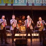 Four male singers perform on stage in grey blazers, red ties, and black trousers, each holding a microphone as they sing in harmony. Behind them, a drummer plays at a kit emblazoned with the Big Girls Don’t Cry logo. Above, a large screen reads “FRANKIE VALLI & THE FOUR SEASONS,” indicating a tribute performance. The stage is simply set with chairs and soft lighting, drawing focus to the vocalists.