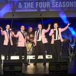 Four male vocalists stand confidently on stage, each wearing a pink blazer with black trousers, white shirt, and black tie, performing in front of a drum kit branded with Big Girls Don’t Cry. They gesture toward the audience, microphones in hand, with a backdrop reading “FRANKIE VALLI & THE FOUR SEASONS,” indicating a tribute performance. Blue spotlights and a seated live band complete the retro showbiz atmosphere.