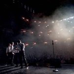 Backstage view of a dramatic theatre scene showing four male performers in white shirts with black braces standing in a dimly lit spotlight haze, facing a large, ornate audience hall filled with spectators. A single microphone stand stands prominently at the front of the stage, silhouetted against the mist and lighting, capturing the anticipation just before a performance begins.