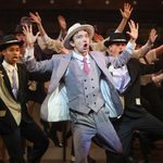A young male performer in light grey three-piece suit and hat is dancing with his arms raised and surrounded by other young male performers in similar costume. Image is from Bugsy Malone, Summer Youth Project 2024.