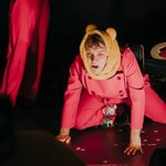 A person in a red jumpsuit and a tan hat with ears is on all fours on a stage, with a shocked expression on their face. The floor is scattered with small, round pieces of confetti.