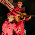 A man in a red jumpsuit and hat plays an acoustic guitar. A woman in a matching outfit is crouched in front of him, looking at the camera and holding up her index finger.