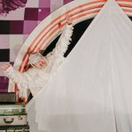 A person in an all-white, Egyptian mummy costume is peeking from behind a large white piece of fabric on a stage. They have a shocked expression and their arms are raised with their hands open. There is a stack of suitcases behind them.