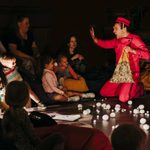 A performer in a red jumpsuit and hat is on their knees, interacting with a small group of adults and children sitting on the floor. The floor is covered in small, white, fluffy spheres.