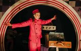 A woman wearing a red jumpsuit and hat stands with her arms outstretched in front of a stage set featuring a large, arched doorway and a pile of vintage suitcases.