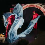 Two performers in red jumpsuits and hats are holding a large, light-blue sheet of fabric. One performer is behind the sheet, and the other is holding it out to the side. The stage has a large arched backdrop with red and white stripes.