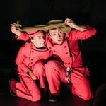 A man and a woman in red jumpsuits and hats are crouched down together. The man is holding a green cloth over both of their heads. They both have surprised expressions on their faces.