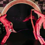 Two performers in red jumpsuits and hats stand on either side of a large arched doorway. They are holding two long, flowing ribbons, one purple and one red, that connect in the middle of the arch.