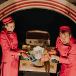 A man and a woman in red jumpsuits and hats are holding up two dolls in front of an open vintage suitcase. There is an arched backdrop behind them.