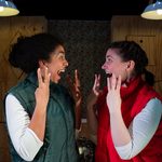 Two performers face each other on a wooden stage set, raising their hands as they react to a surprising discovery.