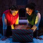 Two performers kneel on the floor, leaning over an open striped gift box lit from within on a festive stage.