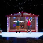 Two performers stand with arms outstretched as artificial snow falls in front of a festive house set decorated with Christmas lights and trees.