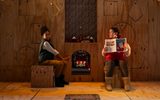 Two people sit beside a glowing fireplace on a festive wooden stage set, with one reading a Christmas book.