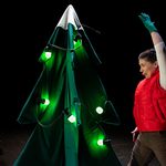 A performer in a red vest raises one arm beside a stylised Christmas tree decorated with glowing lights on a dark stage.
