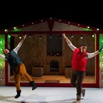 Two performers strike dramatic poses with outstretched arms in front of a house-shaped stage set decorated with Christmas lights.