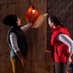 Two people examine a glowing light hidden behind a wooden panel.