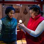 Two performers stand on a festive stage set, looking at a glowing Christmas light bulb held between them.